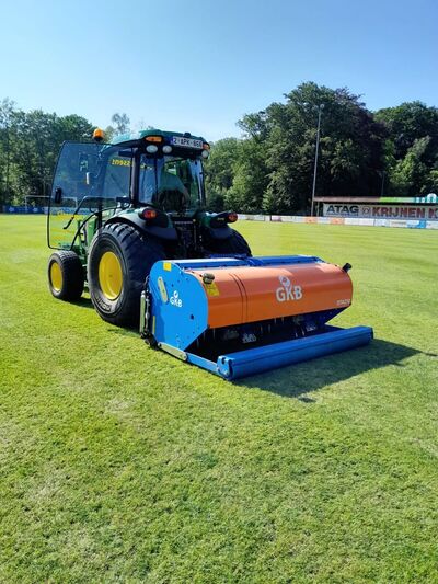 John Deere smalspoortractor met blauw-oranje GKB-bezandingsmachine op een voetbalveld