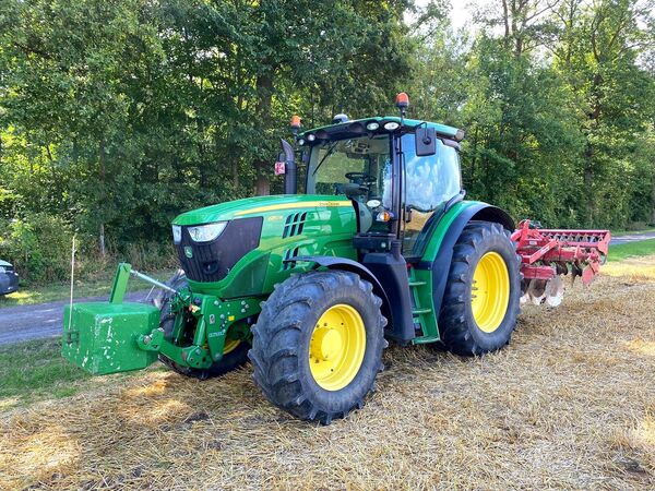 John Deere 6150R met frontgewicht en rode schijveneg langs een bosrand